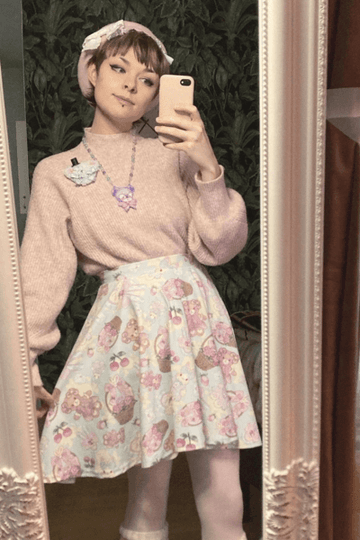 A stylish individual posing in front of a mirror, wearing a pastel sweater and playful patterned skirt.